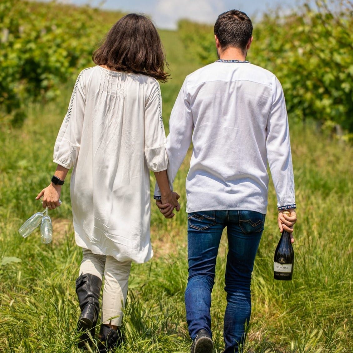 Cuplu d etineri care se tin de mana se plimba prin vie cu o sticla de vin si 2 pahare. Crame din România