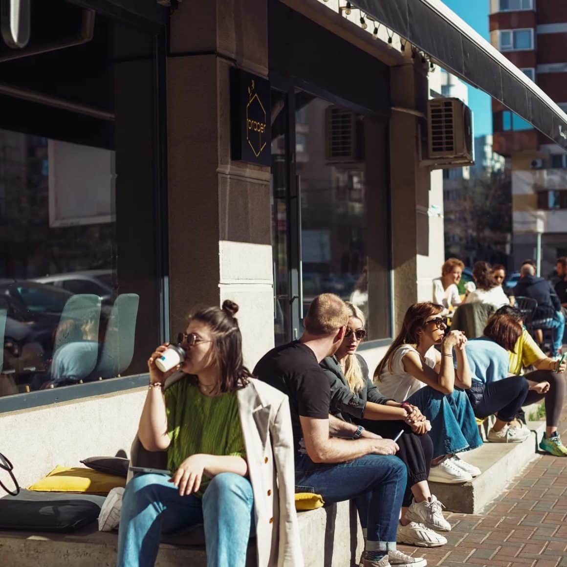 mai multi oameni asezati pe bancutele din fata patiseriei Grain Trip, vorbesc, beau cafea si stau la soare. Cele mai bune deserturi din București