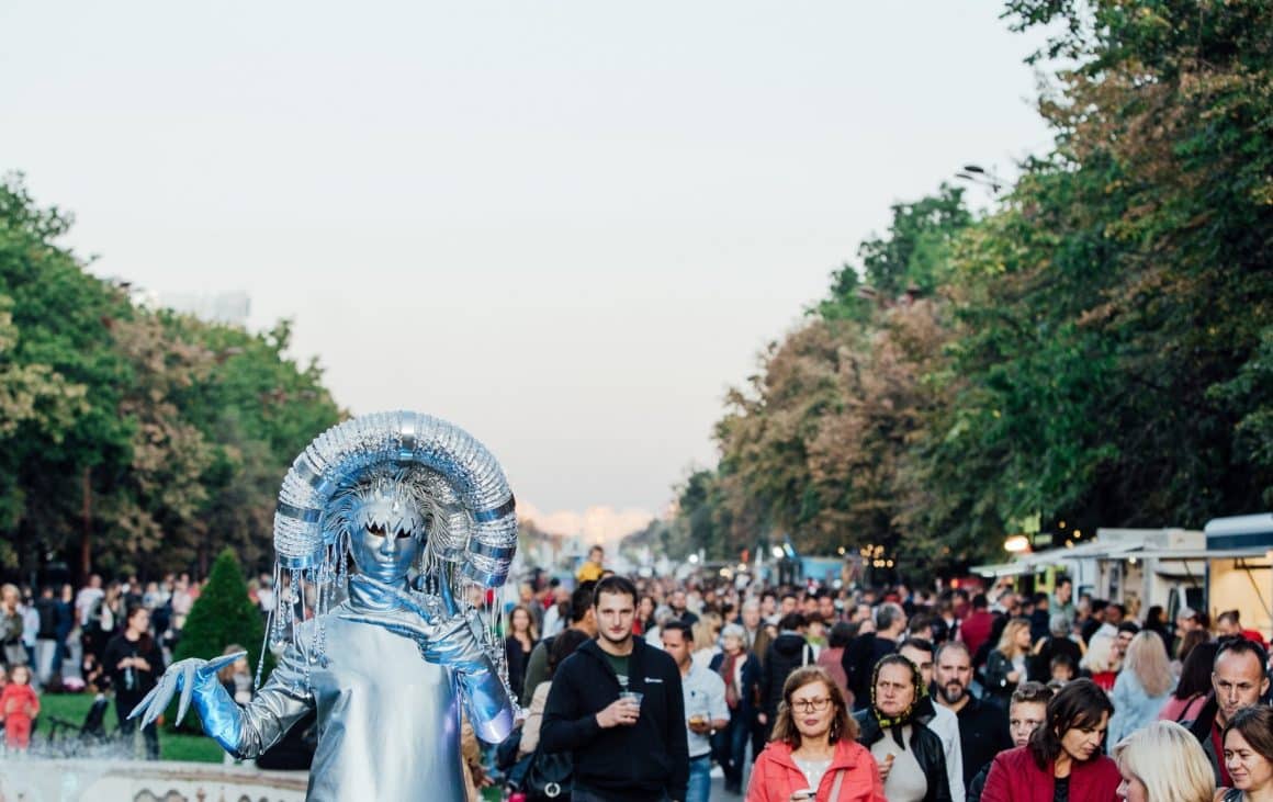 Zilele Bucureștiului. București 365. Evenimente în București de zilele Capitalei. Mai multi oameni la plimbare in aer liber, la un festival, dintre care unul costumat ca o statuetă.