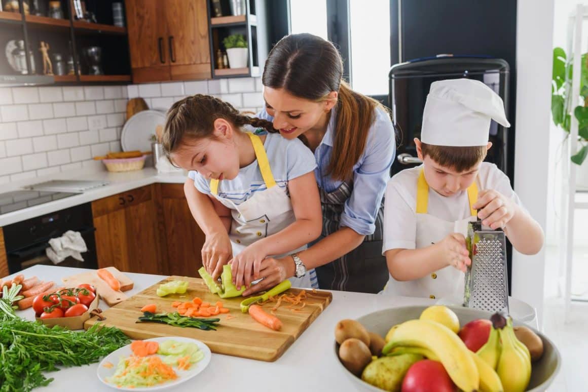 o mama cu doi copii, fata si baiat, prepara mancare in bucatarie. le arata cum sa taie legume si sa le dea pe razatoare