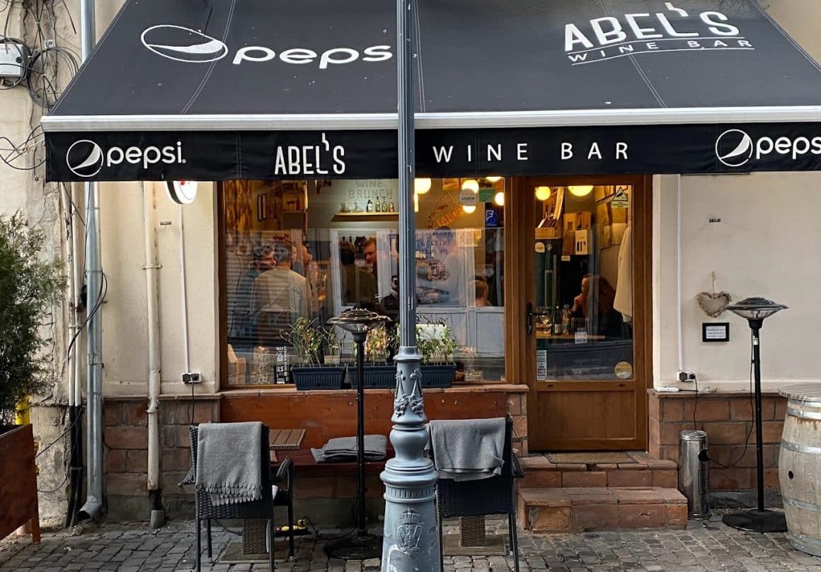 terasa mica de la abel's wine bars București, cu copertina neagra si o masa afara, iar la interior oameni care iau masa. wine bars București