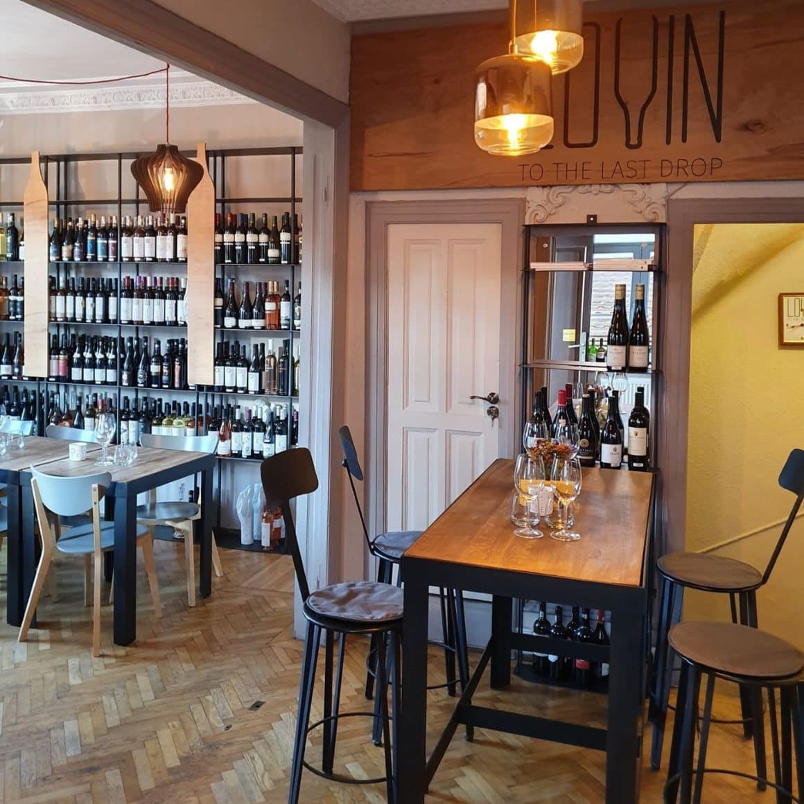 interiorul de la Lovin Wine Bar, cu o masa inalta de bar si 4 scaune, si o alta masa joasa asezata in dreptul unor rafturi de perete, pline cu sticle de vin