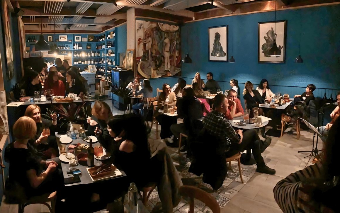 Mai multi oameni iau masa seara la Mosto Natural WineBar & Bistró, intr-o incapere cu pereti albastru inchis, cu multe tablouri si lumina ambientala, iar in fundal barul plin cu sticle de vin. Top wine bars București