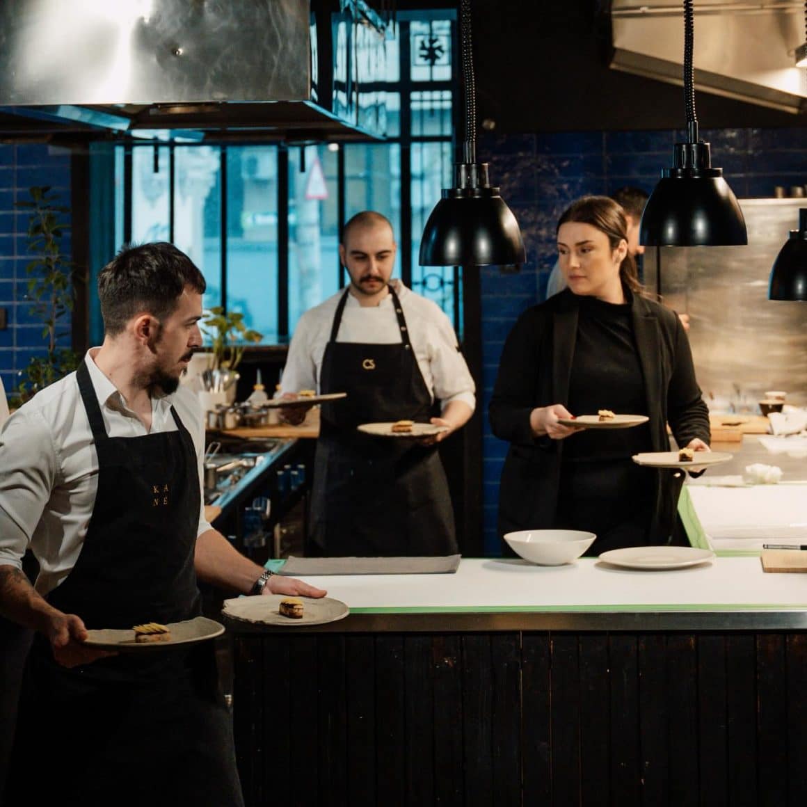 echipa de bucatari de la restaurant Kane Bucuresti, se pregateste sa serveasca farfurii la mese. mai multi oameni fotografiati de la spate, iar masa la Kaiamo București, iar in fundal echipa din bucatarie. Restaurante de fine dining București 