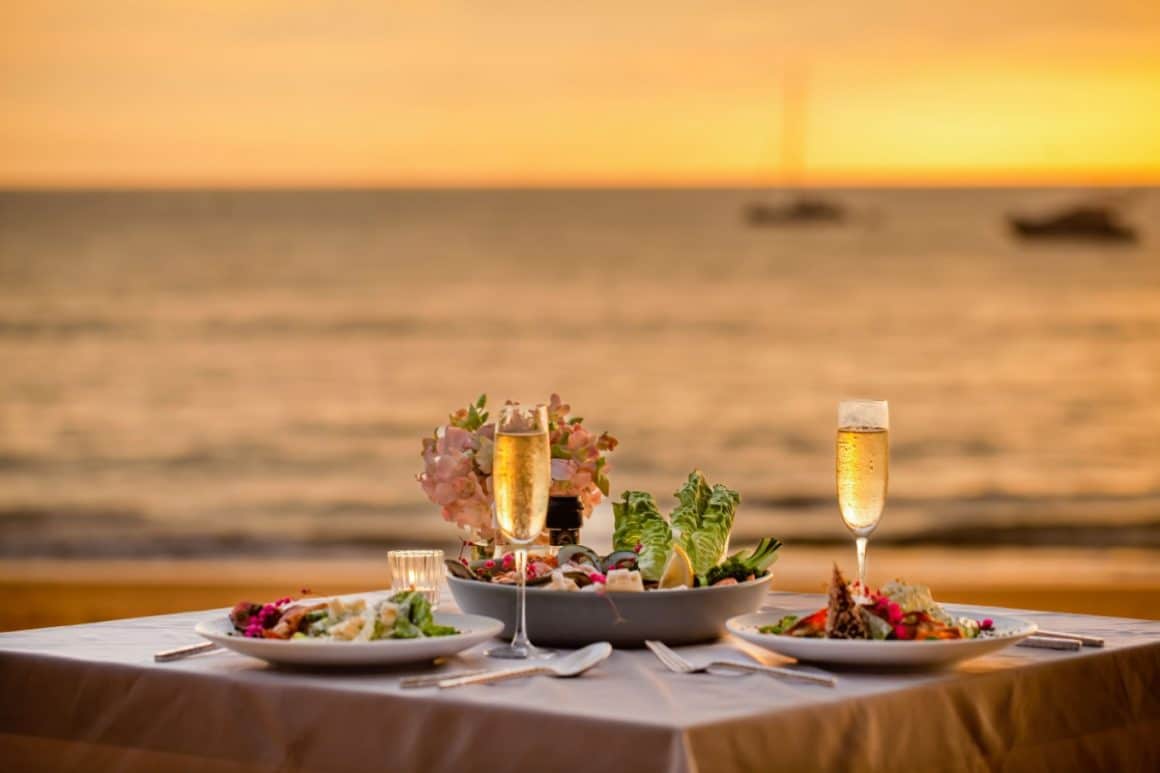 masa la malul marii, fotografiata la apus, cu 2 pahare de vin, un bol mare de salata si 2 farfurii cu mancare. Restaurante Mangalia