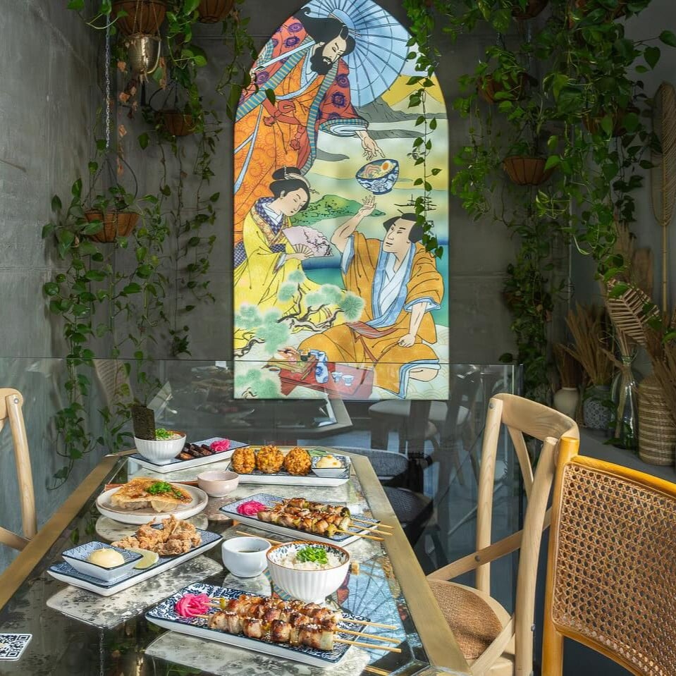 Masa cu preparate japoneze servite la Sanki Ramen Bar, loc din București cunoscut pentru ramen și small plates, iar in fundal un geam vitrat, cu scene japoneze.