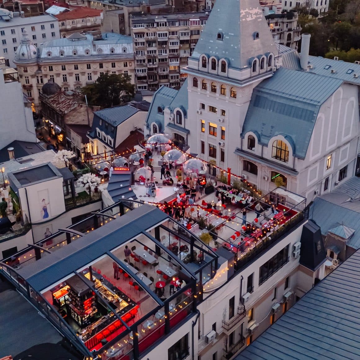 Imagine fotografiata cu drona, de sus, asupra Bartini, cu terasa plina de oameni, seara, si in fundal imagine panoramică asupra capitalei, iar in fundal casa Poporului. Rooftop bars Bucurelști