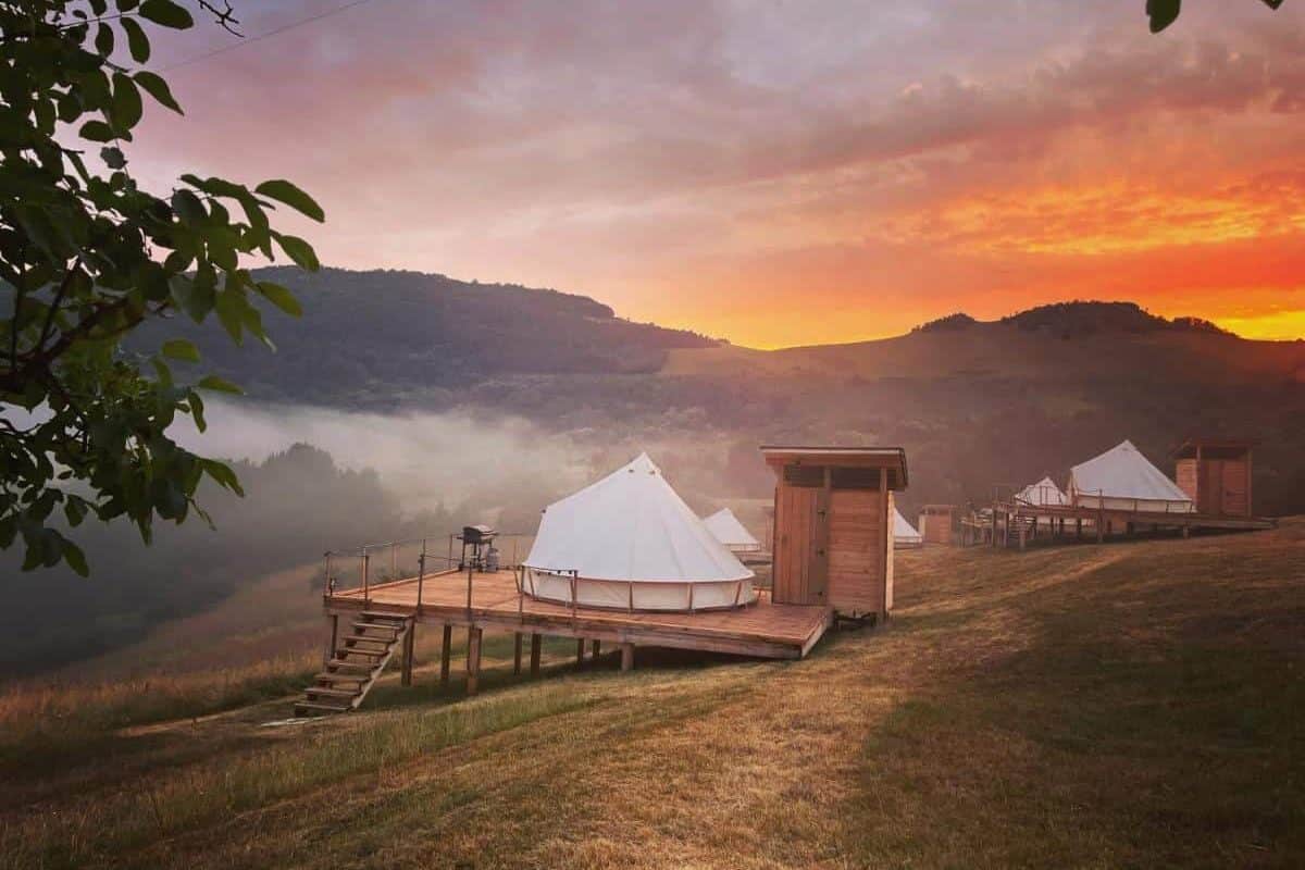 corturi de glamping rasfirate pe o pajiste, amplasate pe structura de lemn, fotografiate seara la apus la munte la The Hundetrs Glamping. Glamping în România