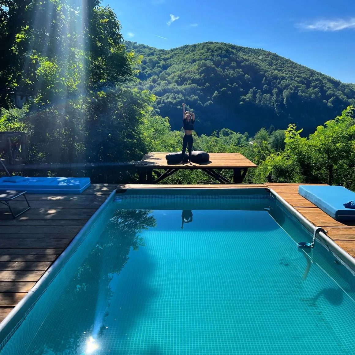 femeie face fitness pe un podet de lemn exterior, in dreptul unei piscina, cu muntii in fundal