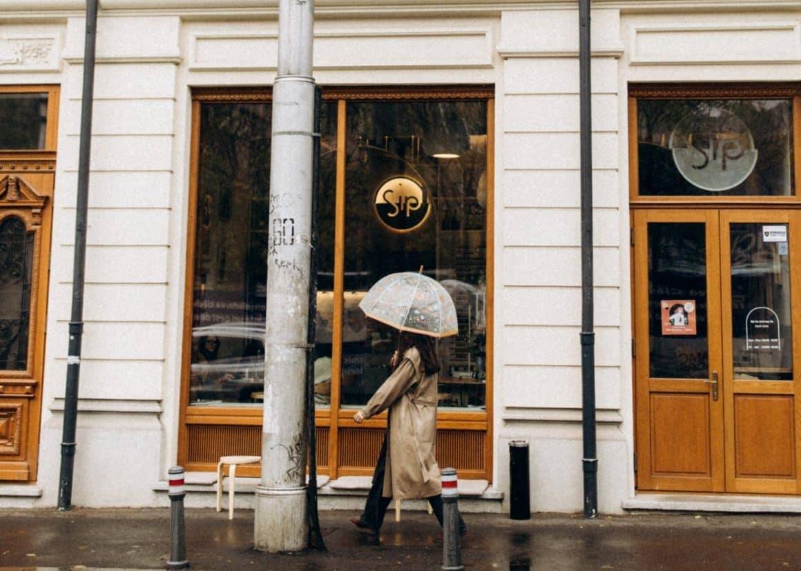 Femeie cu pardesiu si umbrela deschisa trece pe strada prin fata cafenelei Sip Coffee & Wine Bar