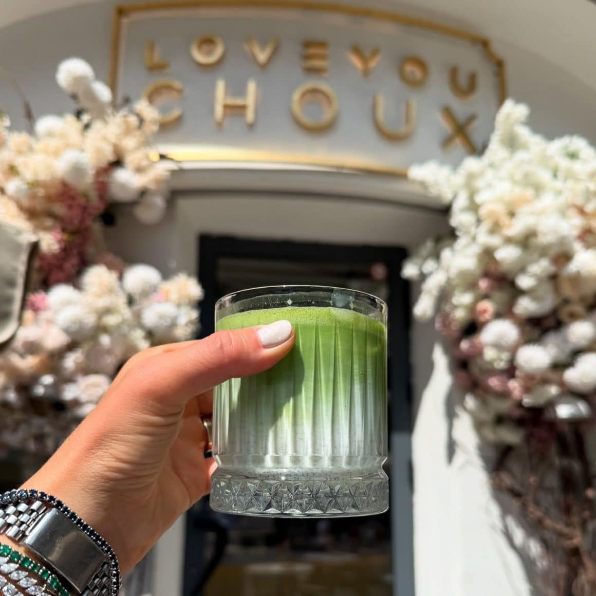 matcha latte rece, servit în pahar scurt, fotografiat în fața intrării cu aranjamente florale.