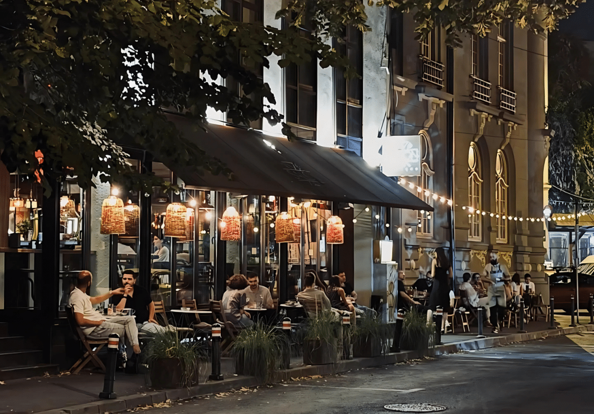 Fațada Salon Golescu seara, lumini calde și mese pline pe trotuar, atmosferă urbană și prietenoasă.