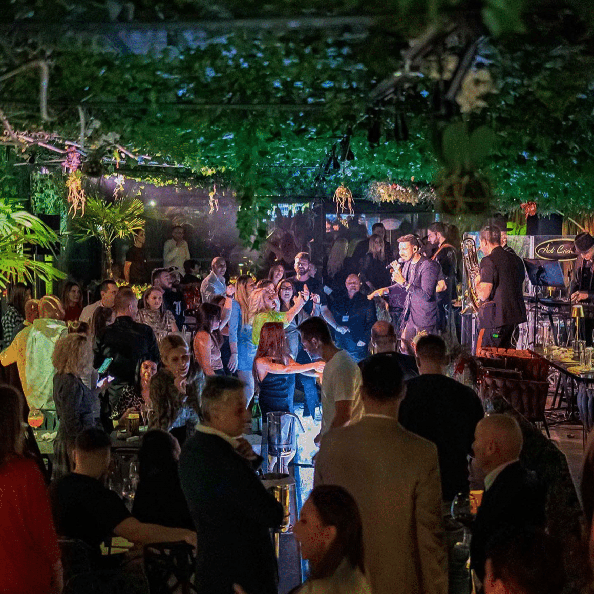 Mai mulți oameni la petrecerea de Crăciun a firmei la Moki București, seara, intr-un spatiu amenajat cu verdeață, și cu band live