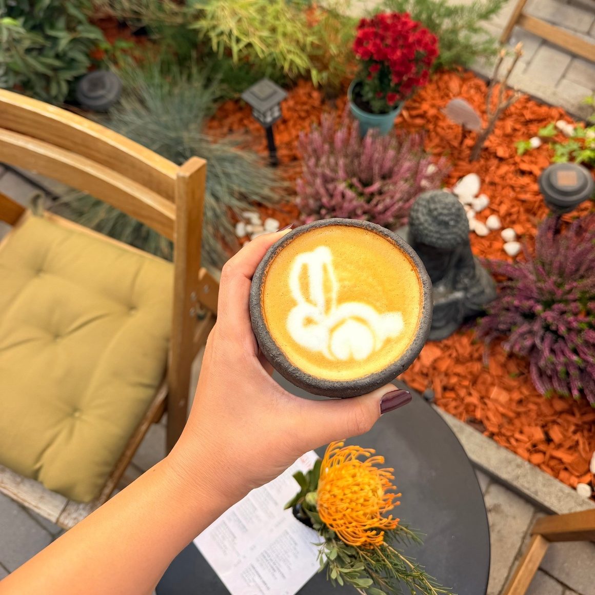 Pumpkin Spice Latte la Ikai, București, servit într-o ceașcă ceramică, cu latte art și vedere spre terasa cu plante decorative.